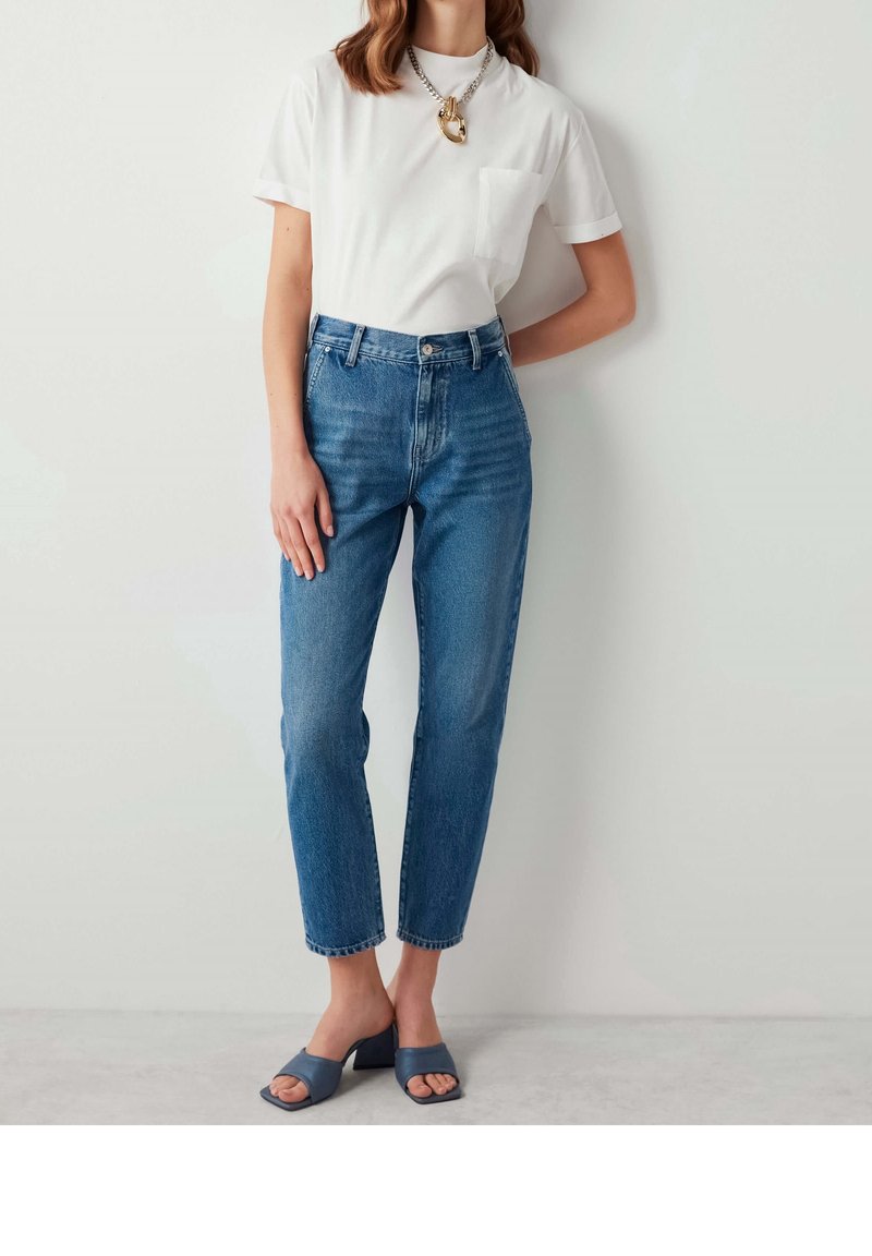 Femme portant un t-shirt blanc à manches courtes, un jean taille haute bleu, un collier chaîne gros maillons or et argent, et des sandales à talons bleues, debout contre un mur blanc.