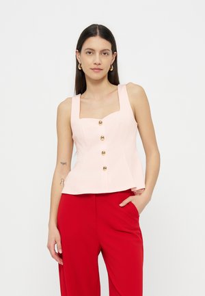 Kobieta z długimi ciemnymi włosami, ubrana w blado różowy top bez rękawów z guzikami i czerwone spodnie, stojąca z jedną ręką w kieszeni.