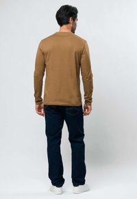 Homme debout de dos portant une chemise marron à manches longues, un jean bleu foncé et des baskets blanches sur un fond gris clair.
