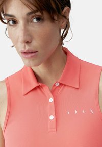 Kora krāsas polo krekls, bez piedurknēm ar apkakli, ar trīs pogu aizdari un baltu logotipu kreisajā krūtī.