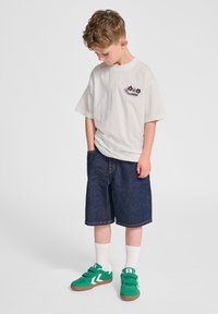 Vit t-shirt med kort ärm och blommönster, kombinerad med mörkblå denimshorts och gröna sneakers, accentuerad med vita strumpor.