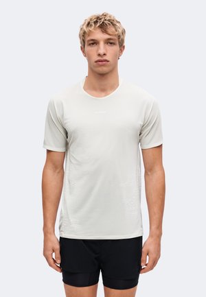 Lyshåret ung mand iført en lysegrå kortærmet skjorte og sorte shorts, stående med front mod kameraet mod en ensfarvet baggrund.