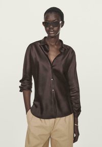 Camicia lunga in satin marrone con vestibilità rilassata, chiusura a bottoni sul davanti, maniche arrotolate, abbinata a pantaloni beige. Gli occhiali da sole aggiungono contrasto.