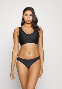 Črni bikini komplet, ki vključuje bralette s širokimi naramnicami v obliko v-izreza ter usklajene nizke hlačke, izdelane iz gladkega in elastičnega materiala.