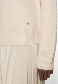 Cremfarbener, gestrickter Pullover mit gerippten Bündchen, der ein kleines goldenes Logo unten links trägt. Weicher Stoff, der zu einem plissierten hellbeigen Rock passt.