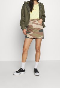 Giacca verde oliva con zip, t-shirt gialla, gonna di jeans con motivo camouflage, calzini bianchi alla caviglia e sneakers nere. Tessuto testurizzato, design informale.