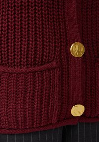 Burgunderfarbener gestrickter Cardigan mit großen goldenen Knöpfen, geripptem Muster und Fronttaschen. Der Stoff wirkt dick und warm.
