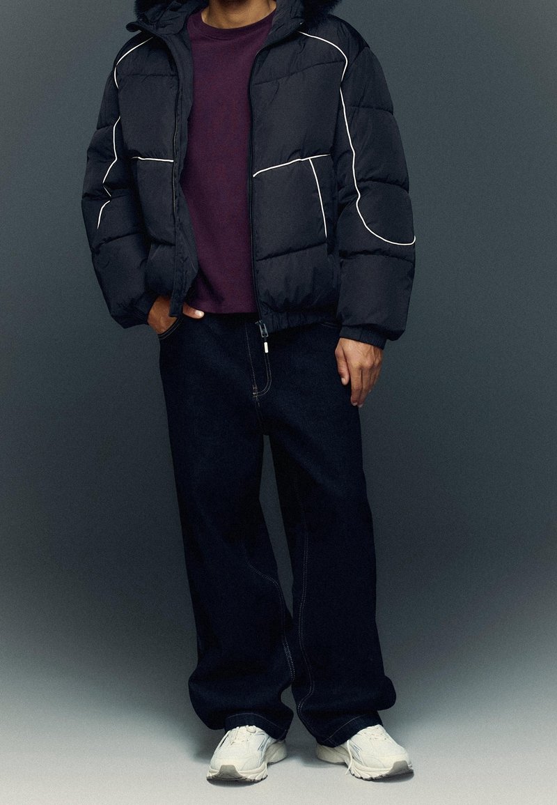 Personne portant une doudoune noire avec des liserés blancs, un jean large foncé, des baskets blanches et une chemise violet foncé, une main dans la poche.