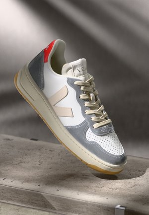 White sneaker with beige "V" logo, gray suede overlays, beige laces, and red heel tab on a concrete surface with soft shadow background.