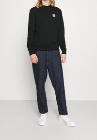 Osoba v černé mikině Lacoste, tmavě modrých volných džínách a bílých vysokých teniskách stojí na pozadí světlého jednobarevného pozadí.