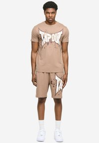 Pješčana pamučna majica kratkih rukava s bijelim i smeđim natpisom 'TAPOUT', uparena s pripadajućim kratkim hlačama istog dizajna. Bijele tenisice zaokružuju izgled.