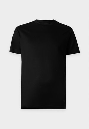 Jeune homme debout portant un t-shirt noir à manches courtes, un jean slim noir et des chaussures en cuir noires sur un fond clair uni.