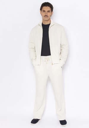 Lichtbeige ritsjack van textuurstof, gecombineerd met bijpassende broek met een trekkoord in de taille. Zwarte shirt eronder, blauwe instappers.
