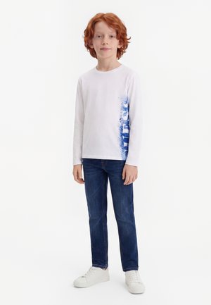 Enfant roux debout, portant un t-shirt blanc à manches longues avec un imprimé bleu sur le côté, un jean bleu et des baskets blanches sur fond blanc.