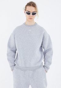 Grauer Sweatshirt aus weichem Material mit Rundhalsausschnitt, tiefen Schultern und dezentem Logo auf der Brust. Trägt man zusammen mit grauen Sweatpants.