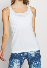 Top bianco senza maniche realizzato in un tessuto leggero e traspirante, con scollatura a barca e spalline sottili, abbinato a leggings blu con motivo.