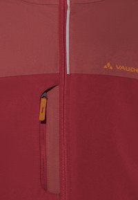 Veste rouge et rouge clair avec une fermeture éclair à l'avant et une poche zippée, présentant une texture lisse et un logo orange sur le côté.