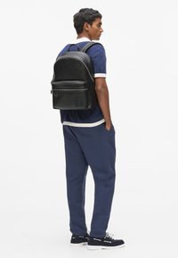 Sac à dos en cuir noir au design arrondi, fermeture éclair argentée, poche avant et logo embossé. Porté avec une tenue bleu marine et des baskets.