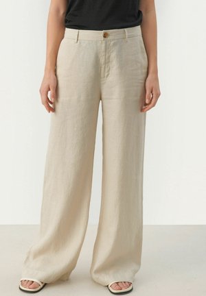 Personne portant un pantalon ample en lin beige avec fermeture par boutons, associé à des sandales blanches ouvertes, debout devant un fond blanc uni.