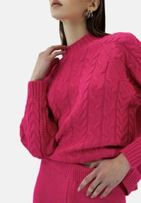 Fuksjowy sweter z dzianiny z wzorem w warkocze, z ściągaczami przy mankietach i dekolcie. Ręka modelki na kołnierzu, zestawiony z pasującą fuksjową spódnicą.