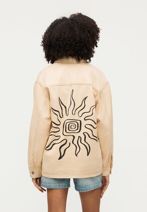 Kobieta z kręconymi włosami, ubrana w beżową kurtkę z czarnym spiralnym wzorem słońca na plecach i niebieskie dżinsowe szorty, stoi tyłem.