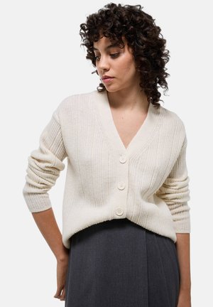 Cardigan en tricot côtelé couleur crème avec un col en V, featuring trois grands boutons et des manches slim légèrement retroussées.