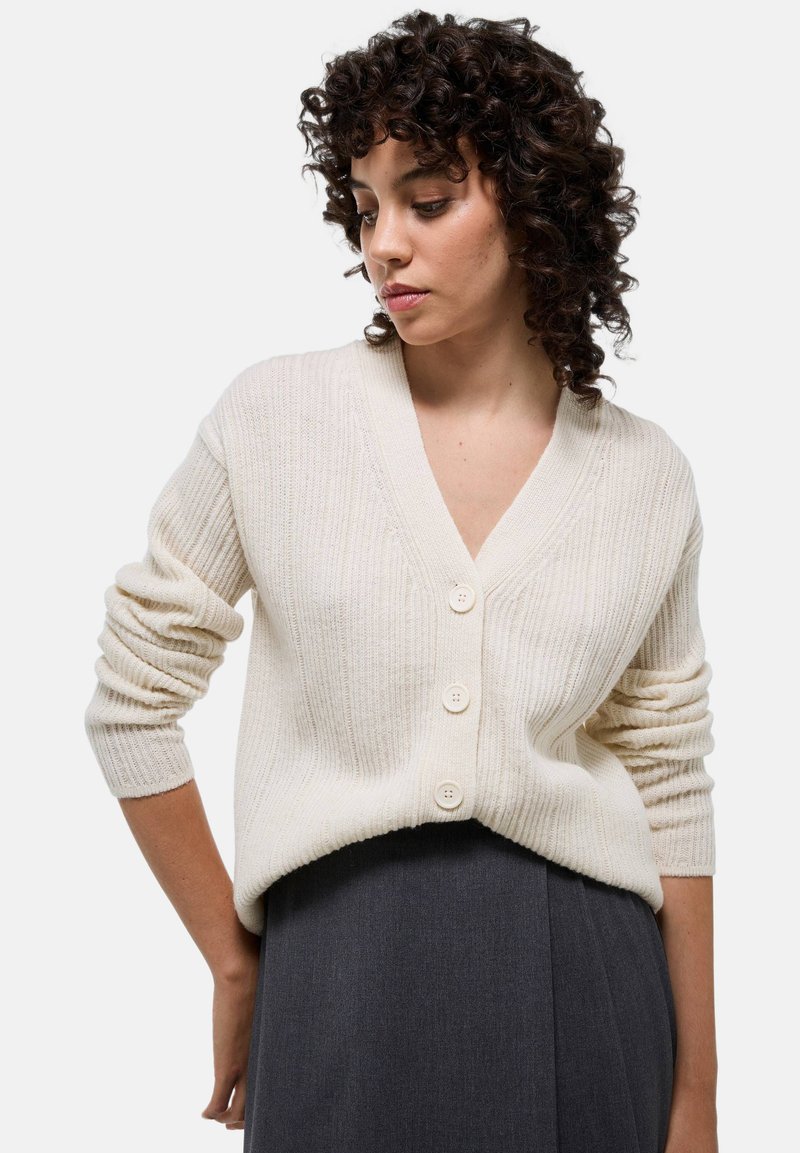Cardigan en tricot côtelé couleur crème avec un col en V, featuring trois grands boutons et des manches slim légèrement retroussées.