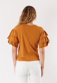 Top giallo senape con maniche a volant a strati, materiale in cotone, scollatura rotonda e vestibilità rilassata, abbinato a pantaloni bianchi slim.