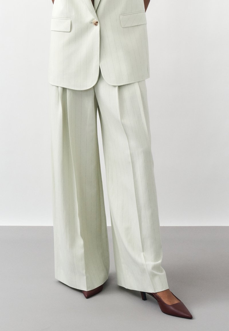 Γυναίκα που φοράει ανοιχτόχρωμο παντελόνι με ρίγες και πλατύ πόδι, συνδυασμένο με σακάκι που ταιριάζει και καφέ μυτερά παπούτσια με τακούνι.