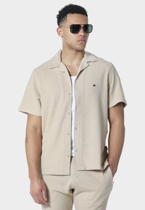 Mężczyzna ubrany w beżową, krótkorękawową koszulę na guziki oraz dopasowane spodnie, z białą podkoszulką i ciemnymi okularami przeciwsłonecznymi, stoi z jedną ręką w kieszeni.