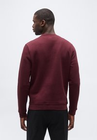 Άνδρας με την πλάτη, φοράει απλό μακρυμάνικο μπλουζάκι σε μπορντό χρώμα και μαύρο παντελόνι, με φόντο ένα απλό λευκό φόντο.