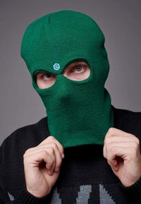 Balaclava verde lavorata a maglia con due aperture per gli occhi, caratterizzata da un logo sulla fronte. Il soggetto indossa un maglione scuro, mentre le mani sistemano la maschera.