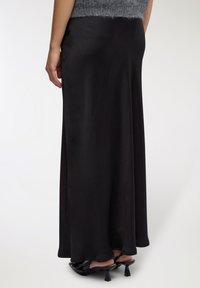 Gonna lunga in satin nero con una texture liscia, caratterizzata da un design fluente e un'ombra delicata. Abbinata a scarpe con tacco nere lucide.