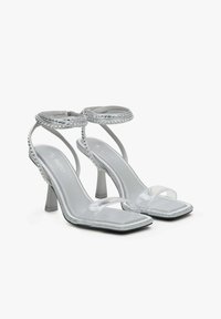 Zilverkleurige sandalen met hoge hakken en doorzichtige banden, vierkante neus en versierd met decoratieve studs langs de randen. Heeft een slanke hak.