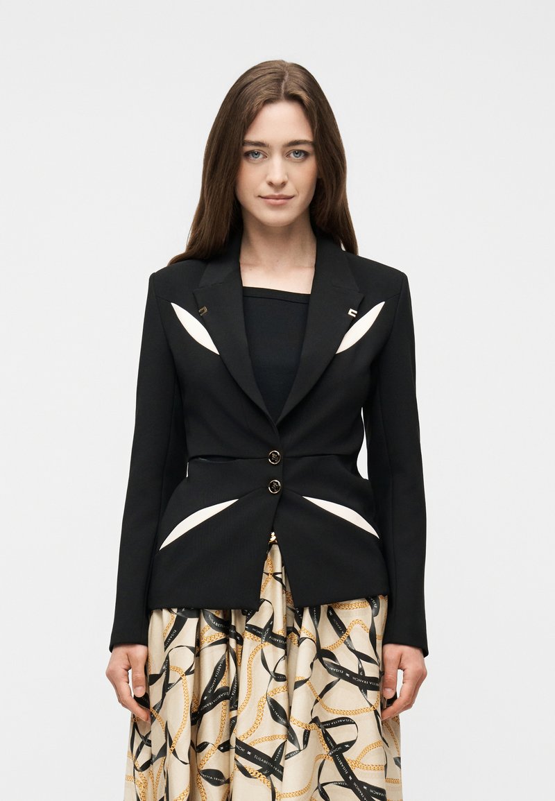 Γυναίκα με μακριά καστανά μαλλιά, φοράει μαύρο σμόκιν με λευκές λεπτομέρειες κοπής και φούστα με σχέδια από χρυσές αλυσίδες και μαύρες κορδέλες.