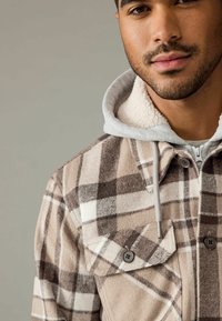 Brun og creme-rutede jakke med en fleece-foret grå hætte, der har to forlommer og knaplukninger. Blød tekstur og varm design.