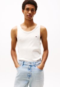 Hvit bomullstank topp med rund hals, med en liten logo på brystet, kombinert med lys blå denimjeans.