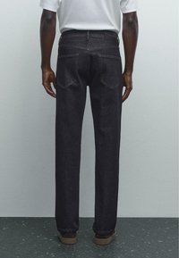 Dark denim jeans with a straight cut, visible back pockets, and a leather label at the waistband. Worn with brown footwear.