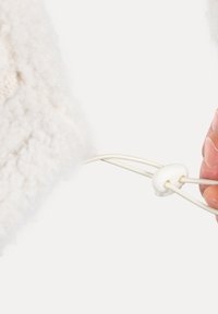 Material blanco y esponjoso con una superficie texturizada; presenta un cordón con un tope blanco que está siendo sostenido por una mano.