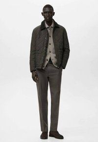 Giacca verde trapuntata con colletto a contrasto, indossata sopra un maglione grigio chiaro, abbinata a pantaloni grigi e scarpe scure.