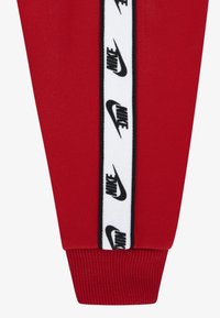 Pantalón deportivo rojo con panel lateral blanco que presenta el logo negro de Nike repetido. Puño elástico en el dobladillo y textura de tela suave en toda la prenda.