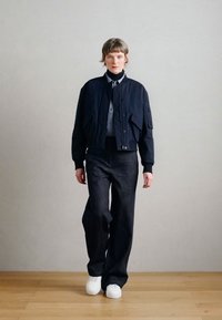 Nila jūras kara flotes bomber jaka ar priekšējām kabatām, kopā ar tumši zilām plašu kāju džinsa biksēm un baltām sporta kurpēm, ar gludu apdari un brīvu piegriezumu.