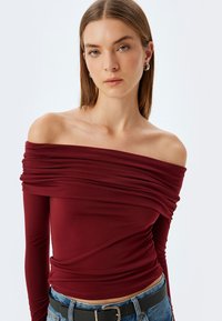 Bordeaux off-shoulder top med samlet tekstur, lange ærmer, tætsiddende design og blødt stof, parret med høj talje denim og et bælte.