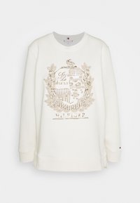 Krämfärgad sweatshirt med en texturerad, metallisk guldemblem och lagerbladiga detaljer. Ribbstickade muddar och nederkant, med sidoflikar för ökad rörelsefrihet.