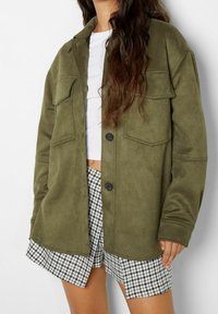 Giacca in tessuto morbido verde oliva, con due tasche sul petto, chiusura a bottoni e vestibilità comoda, abbinata a pantaloni corti in bianco e nero a motivi houndstooth.