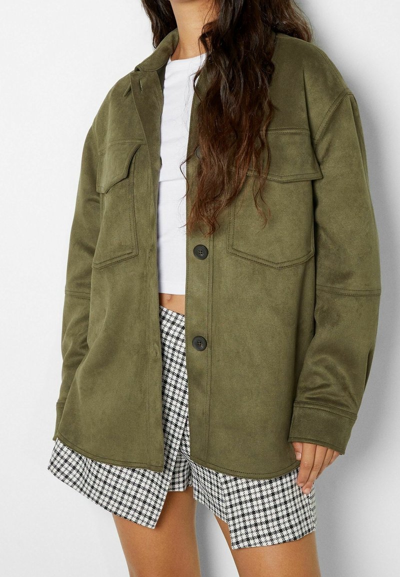 Giacca in tessuto morbido verde oliva, con due tasche sul petto, chiusura a bottoni e vestibilità comoda, abbinata a pantaloni corti in bianco e nero a motivi houndstooth.