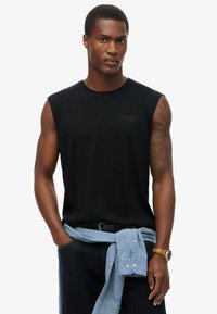 Camiseta sin mangas negra de algodón, con cuello redondo y un logotipo sutil en el pecho. Combinada con pantalones de mezclilla azules y una camisa celeste atada en la cintura.