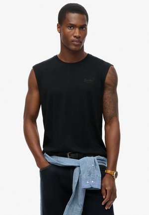 Mężczyzna z tatuażem na prawej ręce, ubrany w czarną koszulkę bez rękawów, jasną dżinsową koszulę zawiązaną w talii, czarne spodnie i brązowy zegarek na nadgarstku.