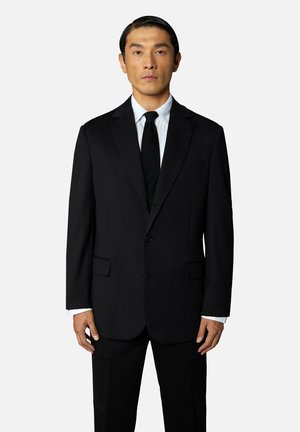 Schwarze Anzugjacke mit Revers, einreihigem Knopfverschluss, zwei vorderen Taschen und passender Hose. Getragen über einem weißen Dress-Shirt und einer schwarzen Krawatte.