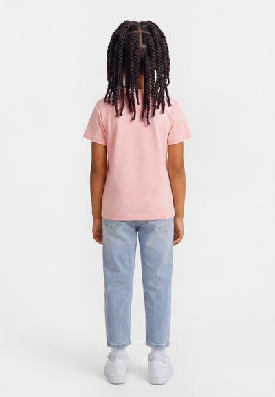 Παιδί με πλεξούδες που φοράει ροζ μπλουζάκι με κοντά μανίκια, ανοιχτό μπλε τζιν, λευκές κάλτσες και λευκά αθλητικά παπούτσια, στέκεται γυρισμένο προς τα πίσω.